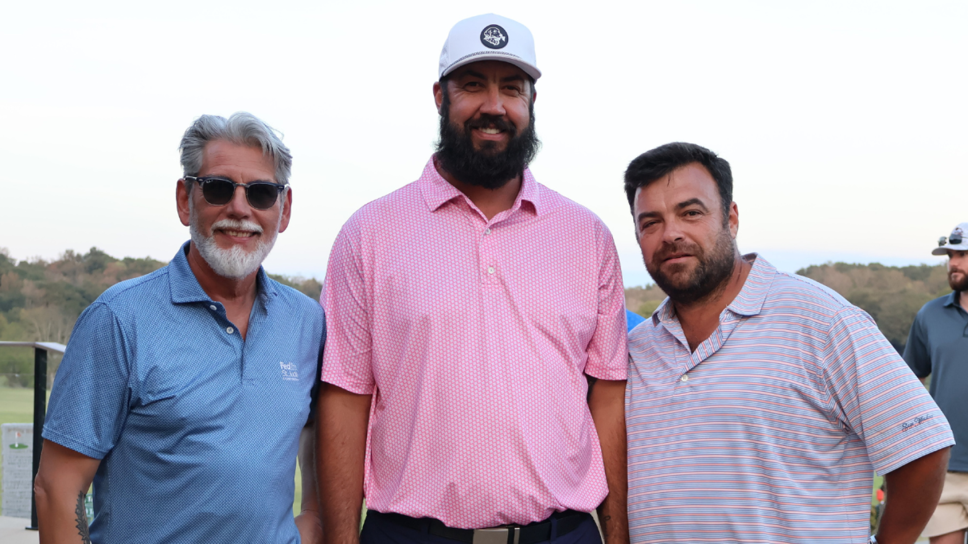 Three men in polo shirts pose together outdoors at a golf course, with trees and a golf cart visible in the background.